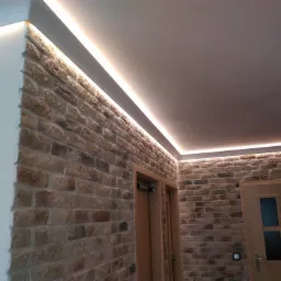 Ściana wyłożona dekoracyjną cegłą z ukrytym oświetleniem LED wzdłuż sufitu, widoczne drzwi w drewnianej ościeżnicy.