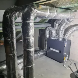 Szare urządzenie rekuperacyjne marki Trinnity z podłączonymi, zaizolowanymi przewodami wentylacyjnymi w pomieszczeniu technicznym, widoczne elementy konstrukcji dachu.