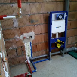 Wnętrze łazienki w trakcie instalacji podtynkowej spłuczki Geberit, widoczne rury doprowadzające wodę i odpływ, ściany z cegły dziurawki, pianka montażowa uszczelniająca przejścia rur.