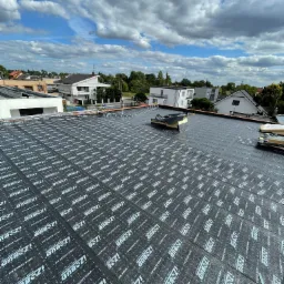 Widok z góry na dach pokryty czarną membraną z białym nadrukiem 'IZOBUD' przed montażem pokrycia, w tle zabudowa jednorodzinna i błękitne niebo z chmurami.