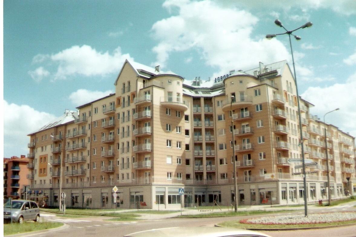 Wielopiętrowy budynek mieszkalny z balkonami, elewacja w odcieniach beżu i brązu, widoczny z poziomu ulicy w słoneczny dzień z lekkim zachmurzeniem.