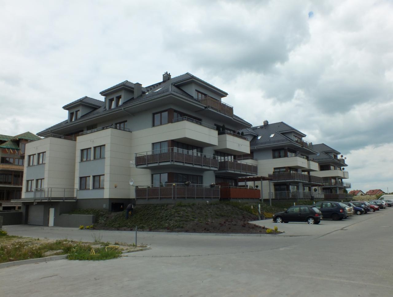 Nowoczesne apartamentowce z balkonami i szarą elewacją, widok z poziomu parkingu w pochmurny dzień.