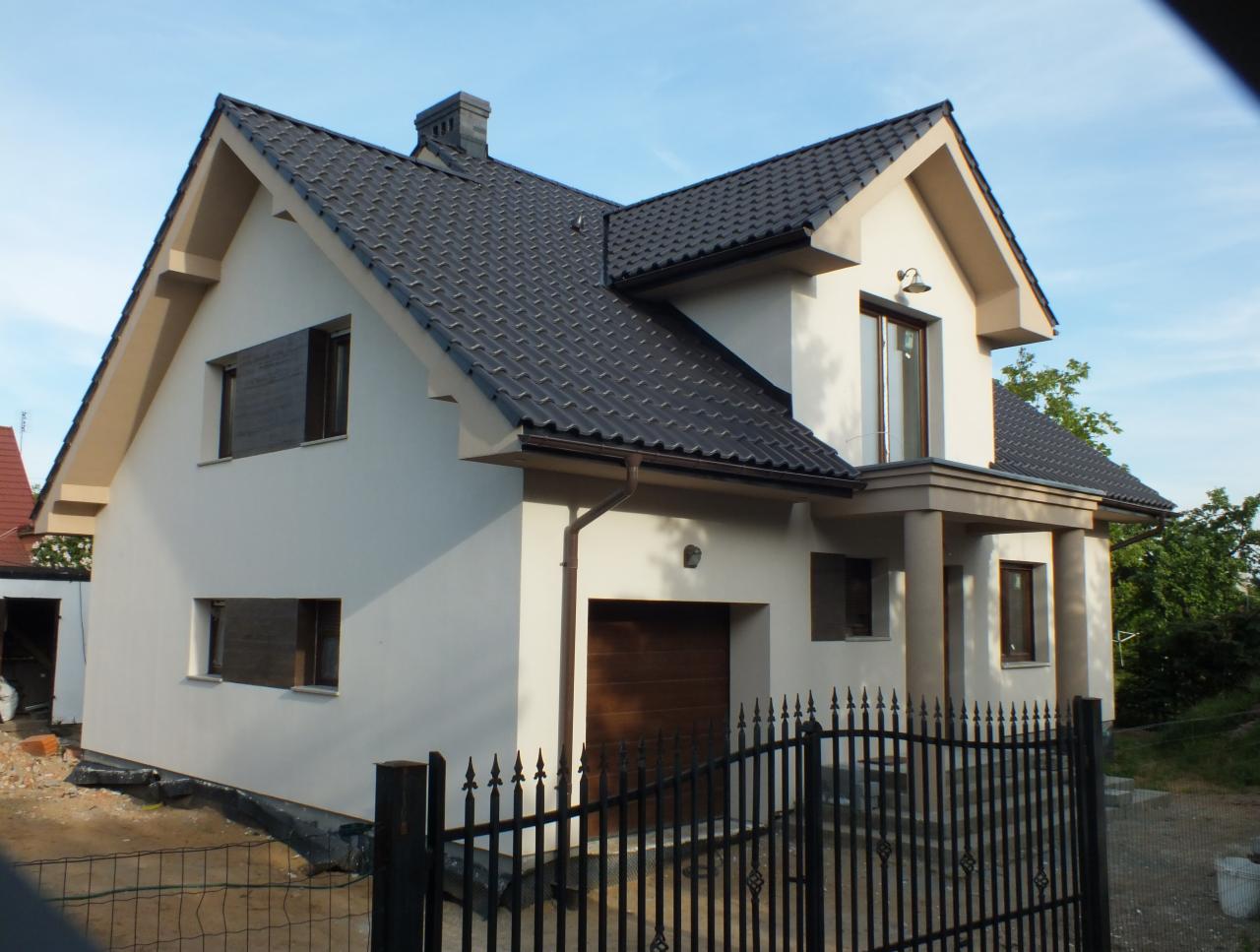 Gotowy dom jednorodzinny z ciemną dachówką, jasną elewacją, garażem i czarnym ogrodzeniem metalowym, widoczny z zewnątrz w słoneczny dzień.