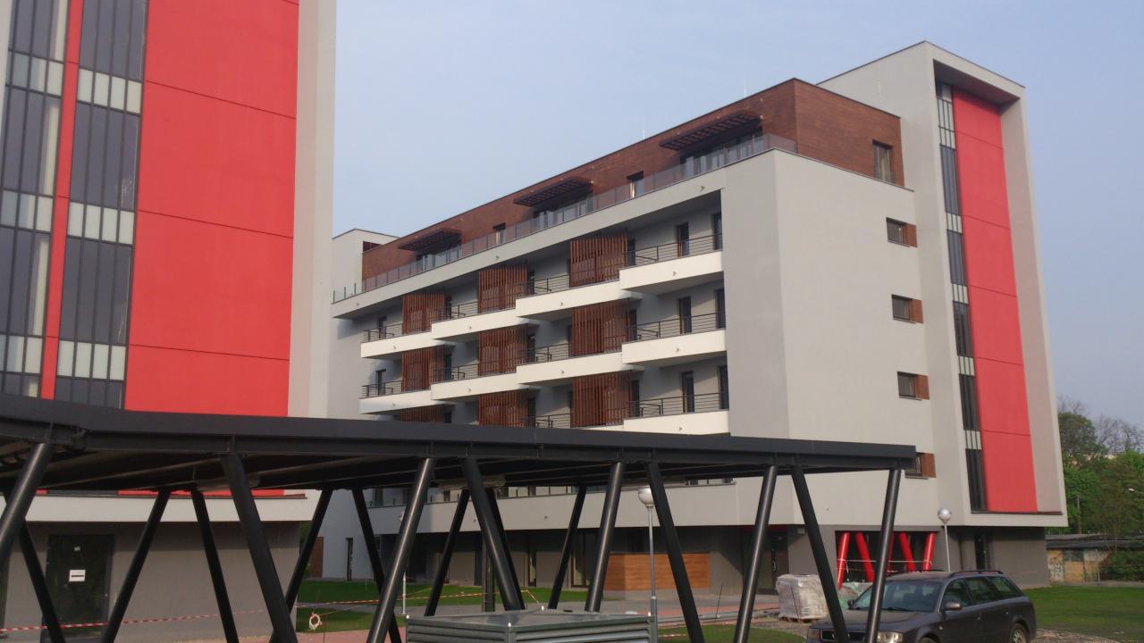 Nowoczesny budynek wielorodzinny z balkonami, elewacją w odcieniach szarości i czerwieni, z drewnianymi elementami na dachu i osłonami balkonów, widziany spod metalowej konstrukcji w pochmurny dzień.