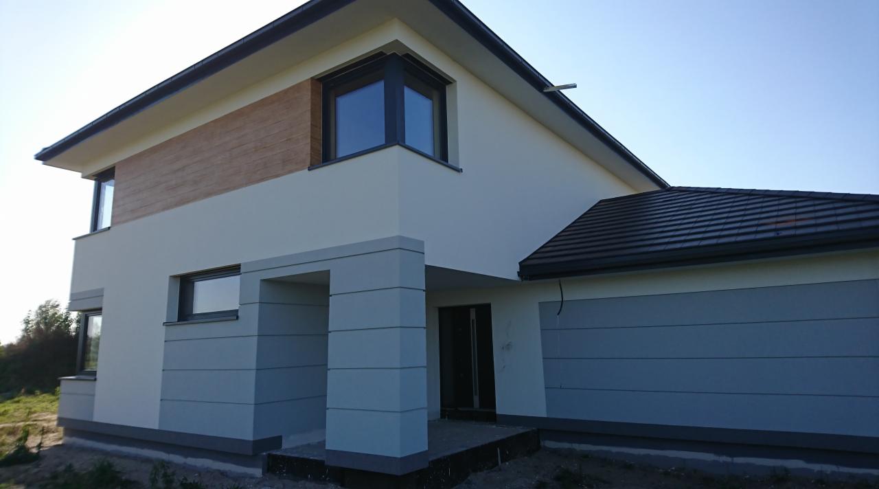 Nowoczesny dom dwukondygnacyjny z elewacją w odcieniach bieli i szarości, fragment drewnianej okładziny na górnej kondygnacji, czarna dachówka, widok z zewnątrz w słoneczny dzień.