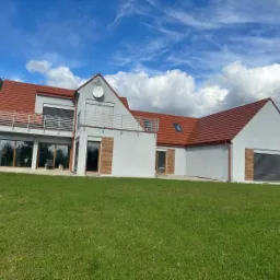 Nowoczesny dom jednorodzinny z czerwonym dachem i szarą elewacją, widok z trawnika w słoneczny dzień z dekoracyjnymi drewnianymi okiennicami.