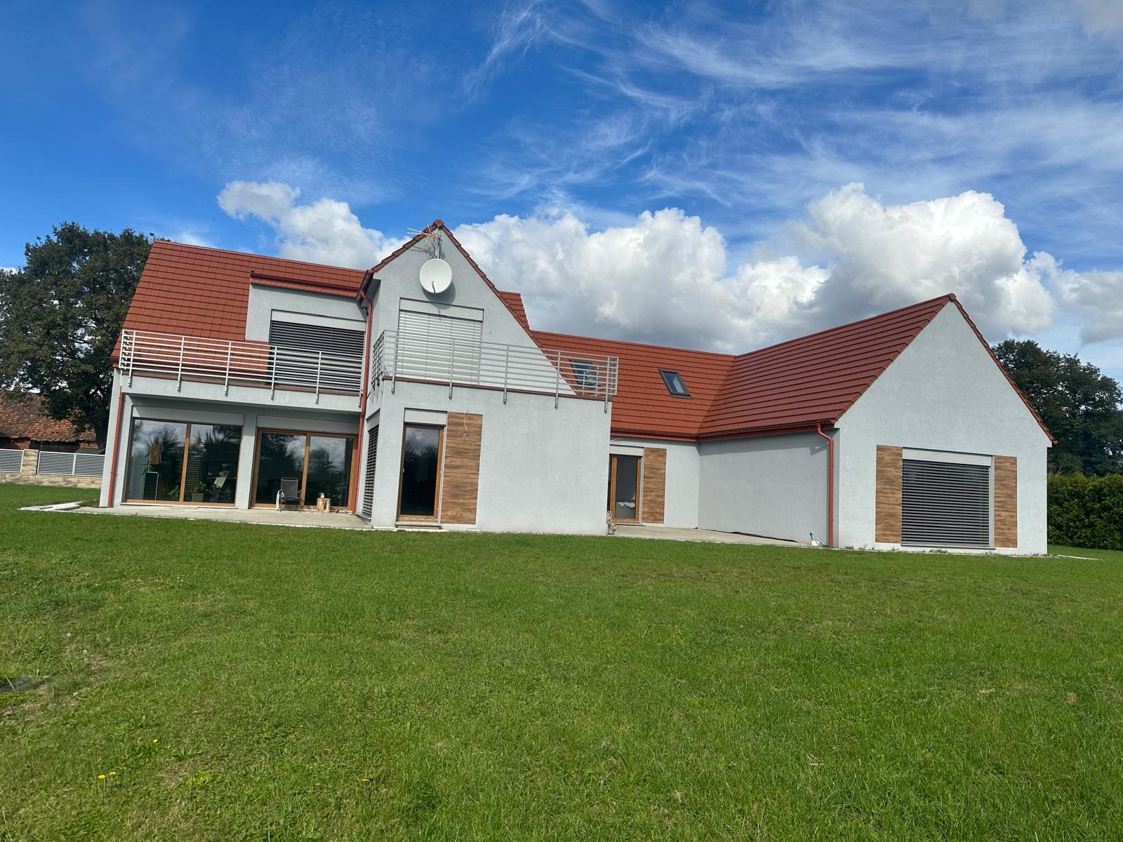 Nowoczesny dom jednorodzinny z czerwonym dachem i szarą elewacją, widok z trawnika w słoneczny dzień z dekoracyjnymi drewnianymi okiennicami.