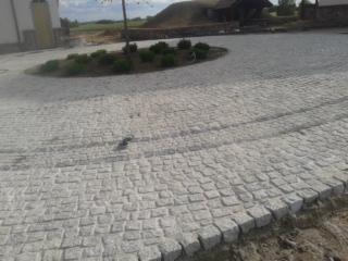 Szara kostka granitowa ułożona na okrągłym podjeździe, widok z góry, zarys budynku w tle, dzień, lekko pochmurne niebo.