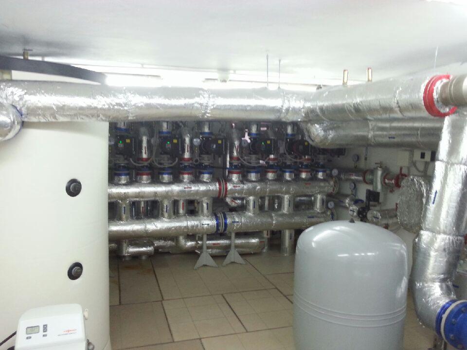 Węzeł cieplny z izolowanymi rurami, zbiornikami i armaturą w pomieszczeniu technicznym. Widoczne liczne zawory i elementy instalacji.