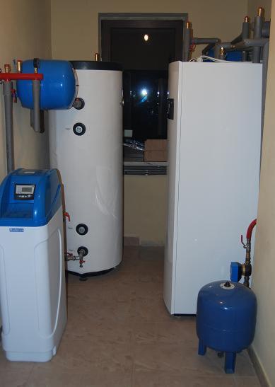 Białe zbiorniki akumulacyjne i niebieskie naczynia przeponowe w kotłowni. Widoczne rury i armatura. Całość zainstalowana w pomieszczeniu z oknem.