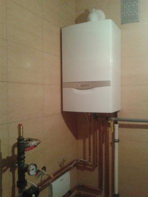 Biały, wiszący piec gazowy marki Vaillant z podłączeniami rur miedzianych w beżowej łazience z płytkami. Widoczne zawory i manometr.