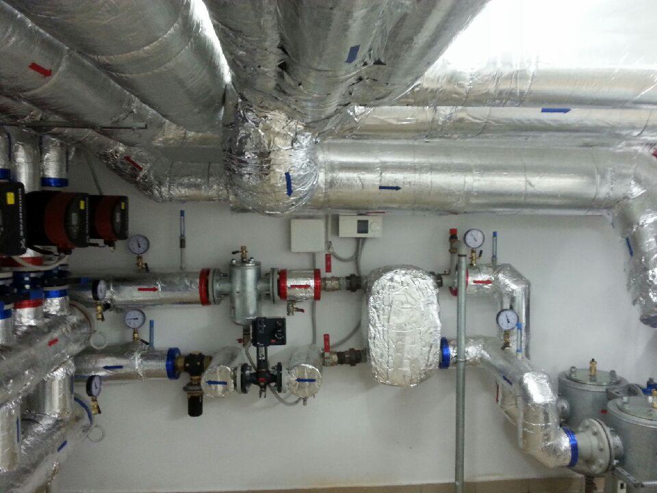 Szczegółowe ujęcie instalacji grzewczej z rurami w srebrnej izolacji, manometrami i zaworami, zamontowanej przy białej ścianie. Widoczne elementy kotłowni.
