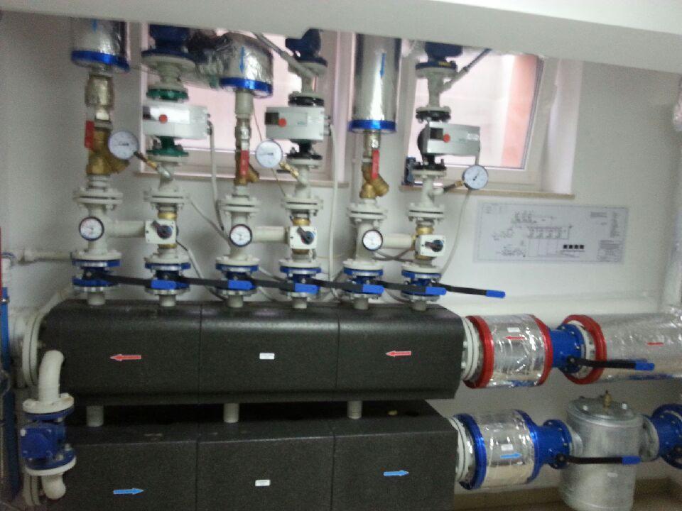 Rozbudowana instalacja grzewcza z czarnymi blokami, rurami, zaworami i manometrami. Schemat na ścianie. Widoczne strzałki wskazujące kierunek przepływu.