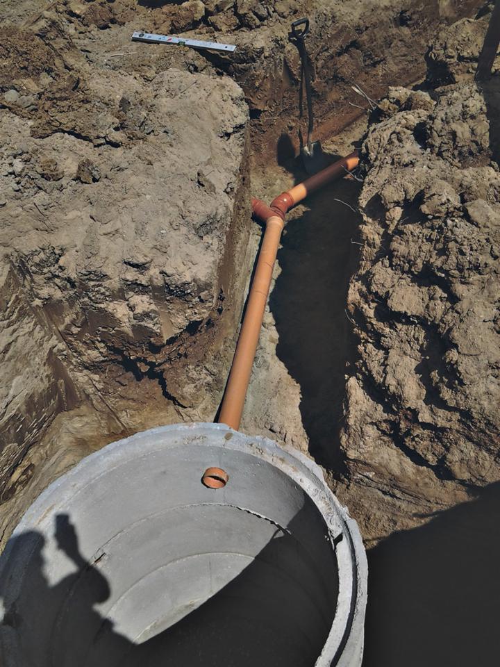 Betonowy krąg studni i pomarańczowa rura w wykopie ziemnym, łopata i poziomica w tle. Ujęcie z góry, ukazujące etap prac instalacyjnych.