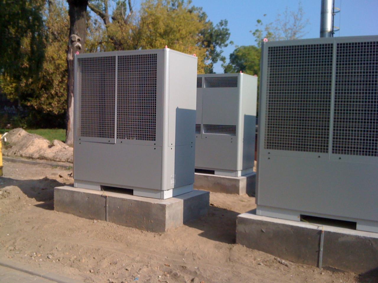 Instalacja trzech szarych pomp ciepła na betonowych fundamentach, z widocznym drzewem i niebieskim niebem w tle. Widok z poziomu gruntu.