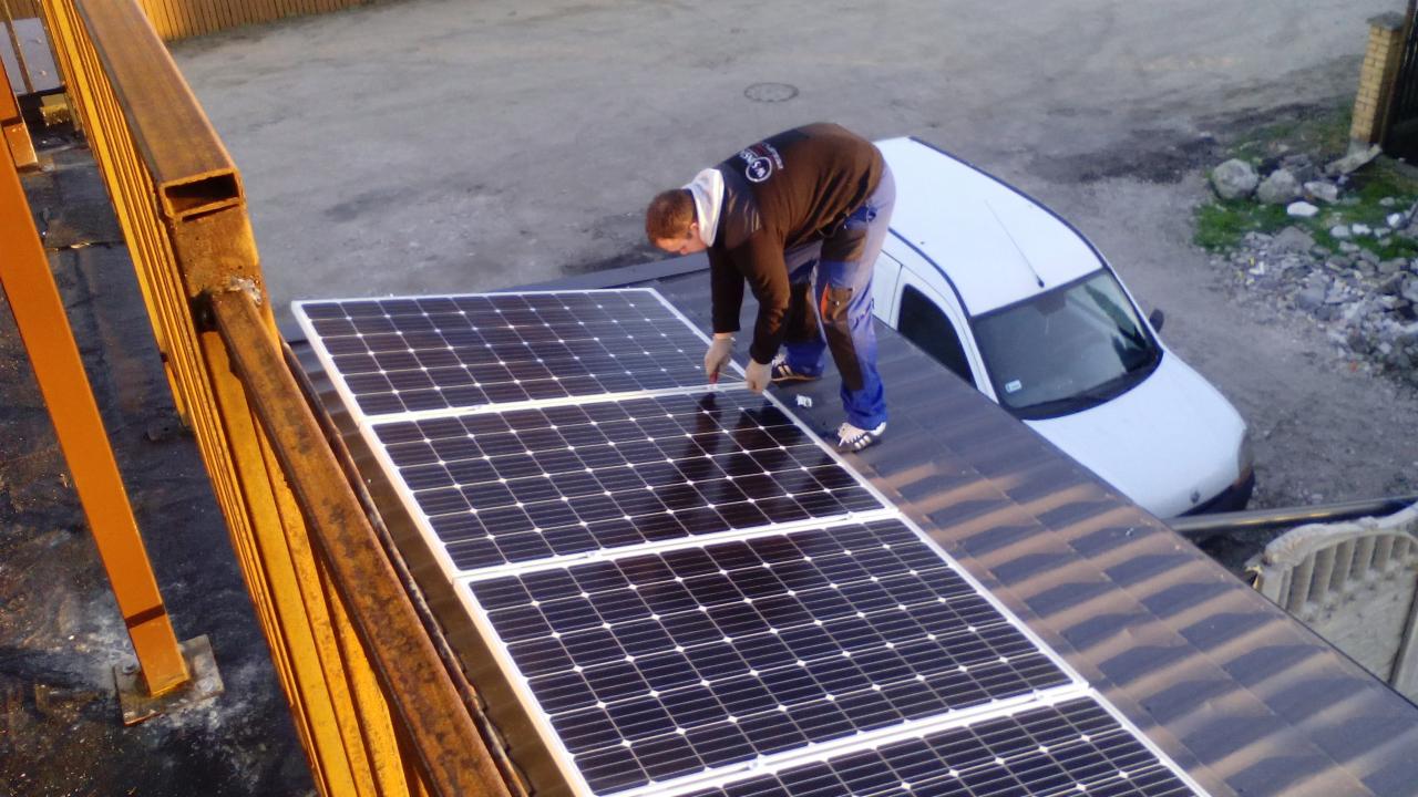 Montaż paneli słonecznych na dachu w Grudziądzu. Instalator w kombinezonie roboczym łączy panele. W tle biały samochód dostawczy i metalowa konstrukcja.