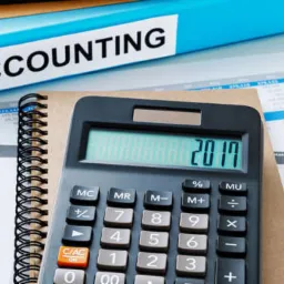 Niebieski segregator z napisem ACCOUNTING, kalkulator z wyświetloną datą 2017, notatnik w twardej oprawie, długopis, okulary i markery na arkuszach kalkulacyjnych.