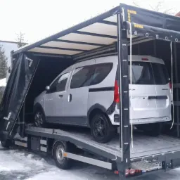 Srebrny samochód dostawczy Dacia Stepway umieszczony na platformie lawety z plandeką w zimowej scenerii.