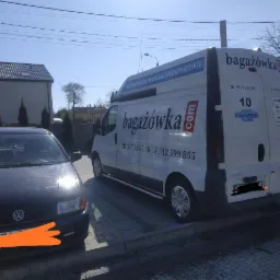 Biały bus firmy transportowej z logo 'bagażówka.com' zaparkowany na kostce brukowej obok czarnego samochodu osobowego, w tle budynek mieszkalny i drzewa.