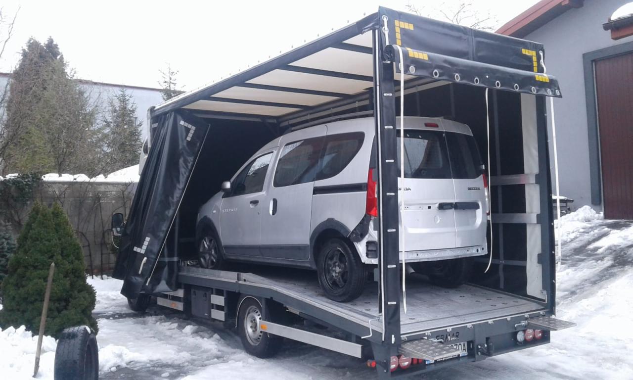 Srebrny samochód dostawczy Dacia Stepway umieszczony na platformie lawety z plandeką w zimowej scenerii.
