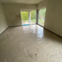 Świeżo wylana, mokra posadzka w jasnym pomieszczeniu z dużymi oknami, widoczne metalowe znaczniki poziomu.