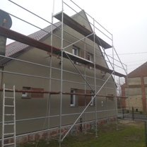 Dom jednorodzinny w trakcie prac elewacyjnych, z widocznym rusztowaniem i fragmentem ocieplenia. Świeżo nałożony tynk w odcieniach beżu. Fragment dachu z brązową dachówką. Widoczne okna. Pochmurne...