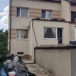 Dwupiętrowy dom z beżową elewacją i brązowymi pasami dekoracyjnymi, widoczne ślady prac malarskich, w tym puste wiadra po farbie i plandeki ochronne, donica z różowymi kwiatami na pierwszym planie.