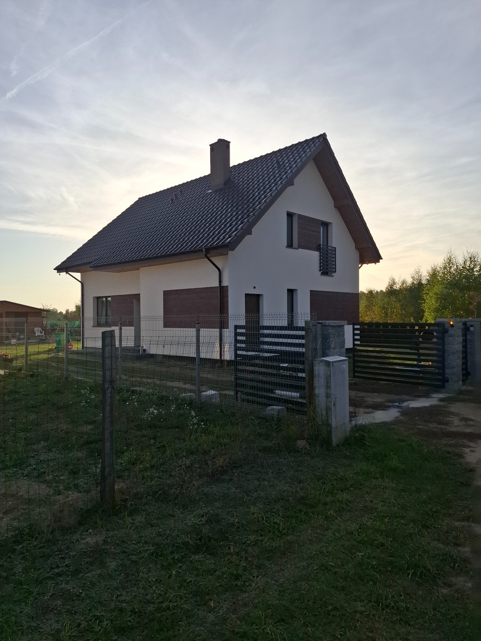 Nowoczesny dom jednorodzinny z białymi ścianami i drewnopodobną elewacją, ogrodzony metalową siatką i bramą wjazdową, w tle zieleń drzew i zachodzące słońce.
