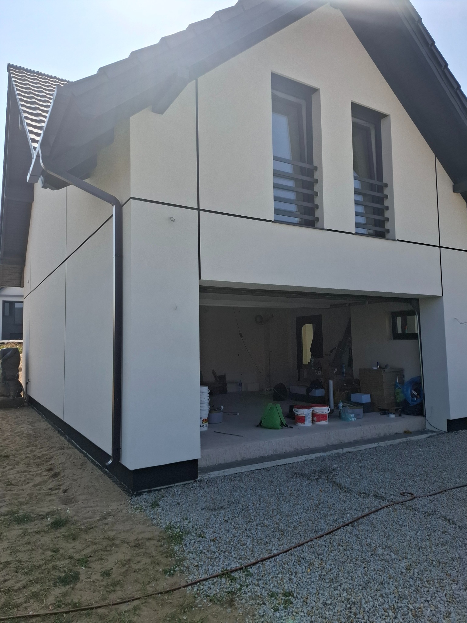 Nowoczesny dom jednorodzinny z wykończoną, jasną elewacją panelową i czarnymi akcentami, widoczne otwarte drzwi garażowe z elementami prac wykończeniowych wewnątrz.