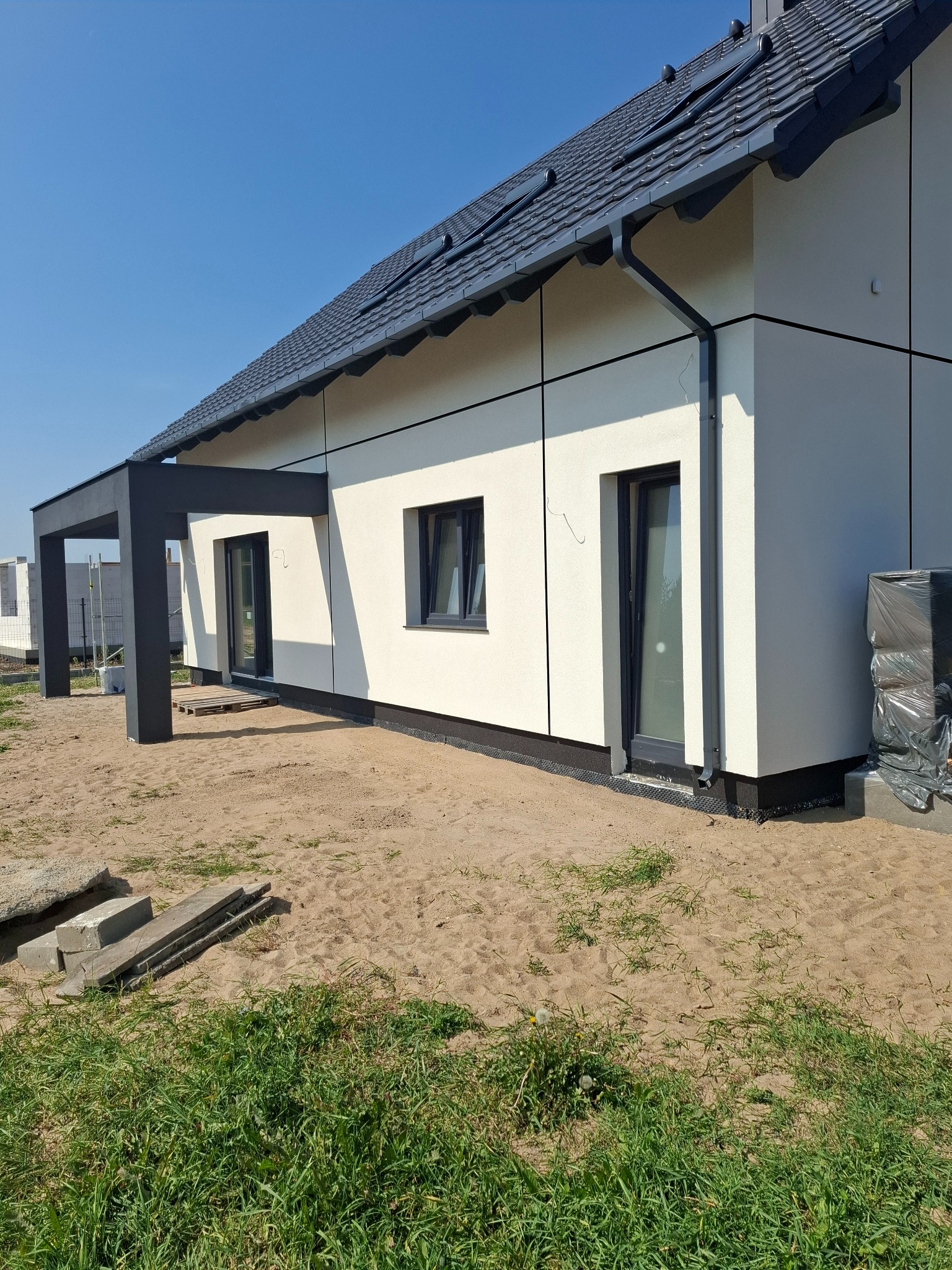 Nowoczesny dom z elewacją w odcieniach bieli i szarości, z widocznymi panelami elewacyjnymi, czarną stolarką okienną i zadaszeniem tarasu. Teren wokół domu w trakcie prac wykończeniowych.