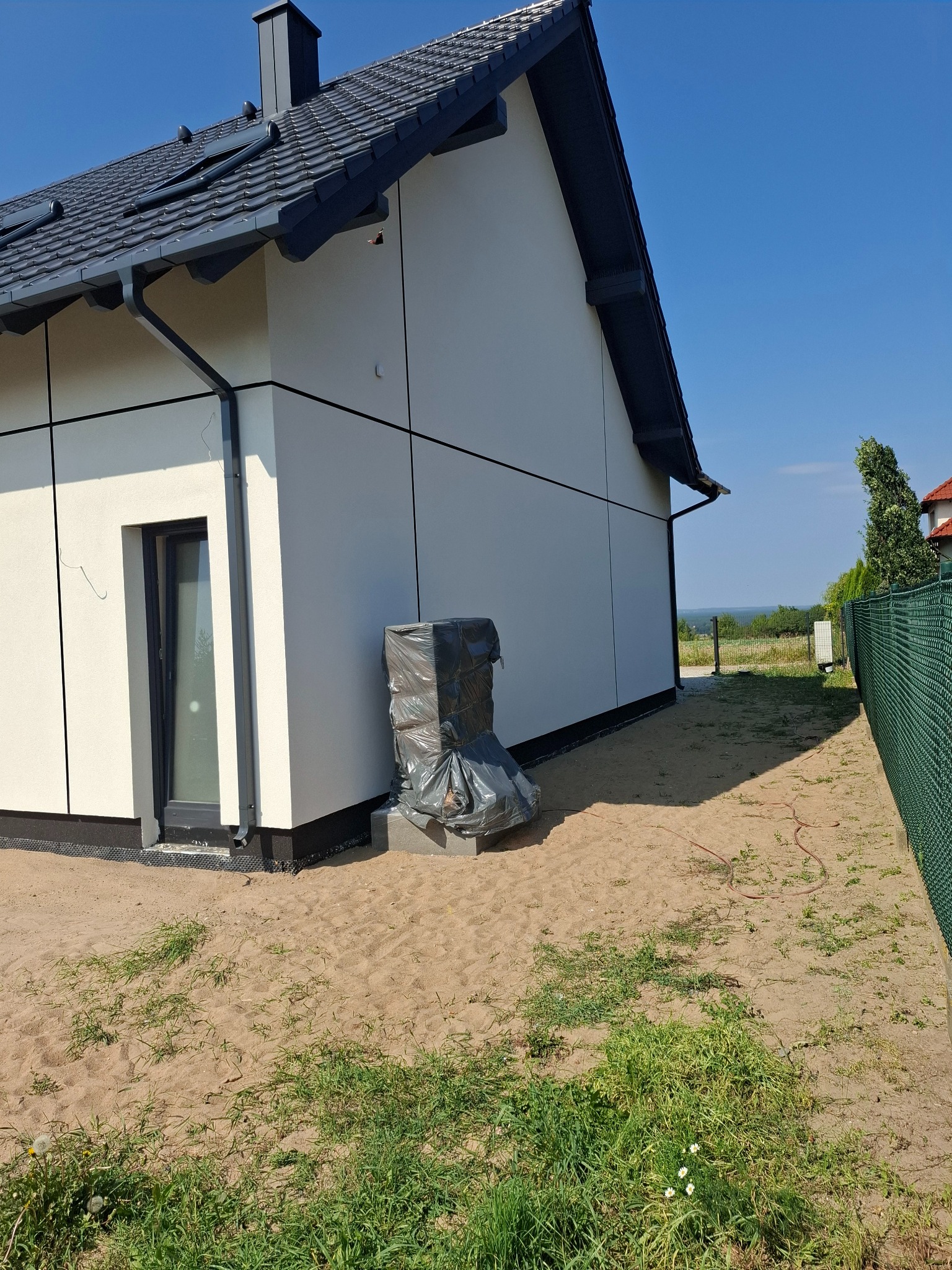 Nowoczesny dom jednorodzinny z jasną elewacją panelową i ciemną stolarką okienną; na ziemi leżą materiały budowlane zabezpieczone folią, widoczna zieleń i ogrodzenie.