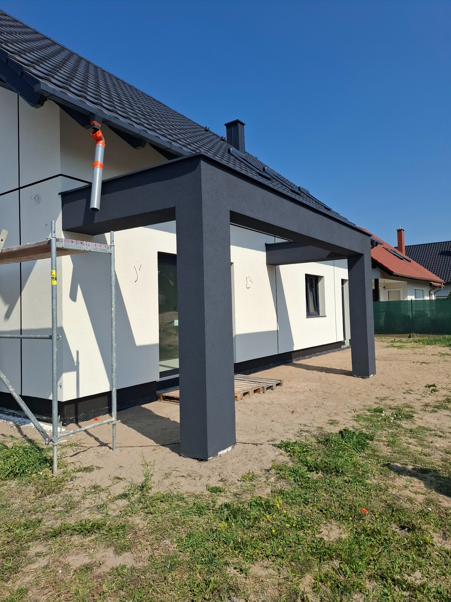 Nowoczesny dom z jasną elewacją i grafitową pergolą przy wejściu, widoczne rusztowanie, teren wokół domu jeszcze niezagospodarowany.