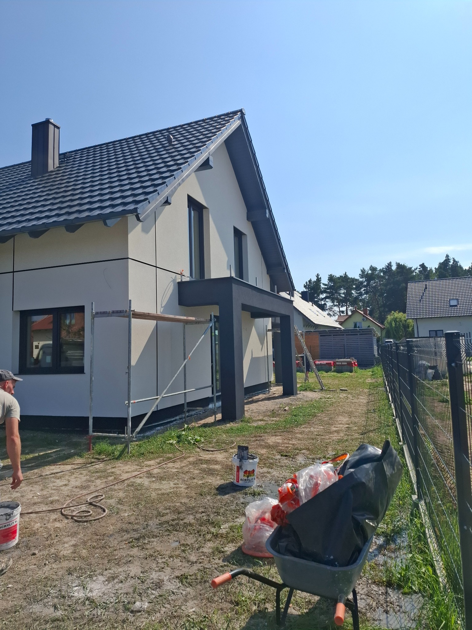 Nowoczesny dom jednorodzinny w trakcie prac elewacyjnych, widoczne rusztowania, wiadro z farbą i taczka z odpadami budowlanymi na posesji.