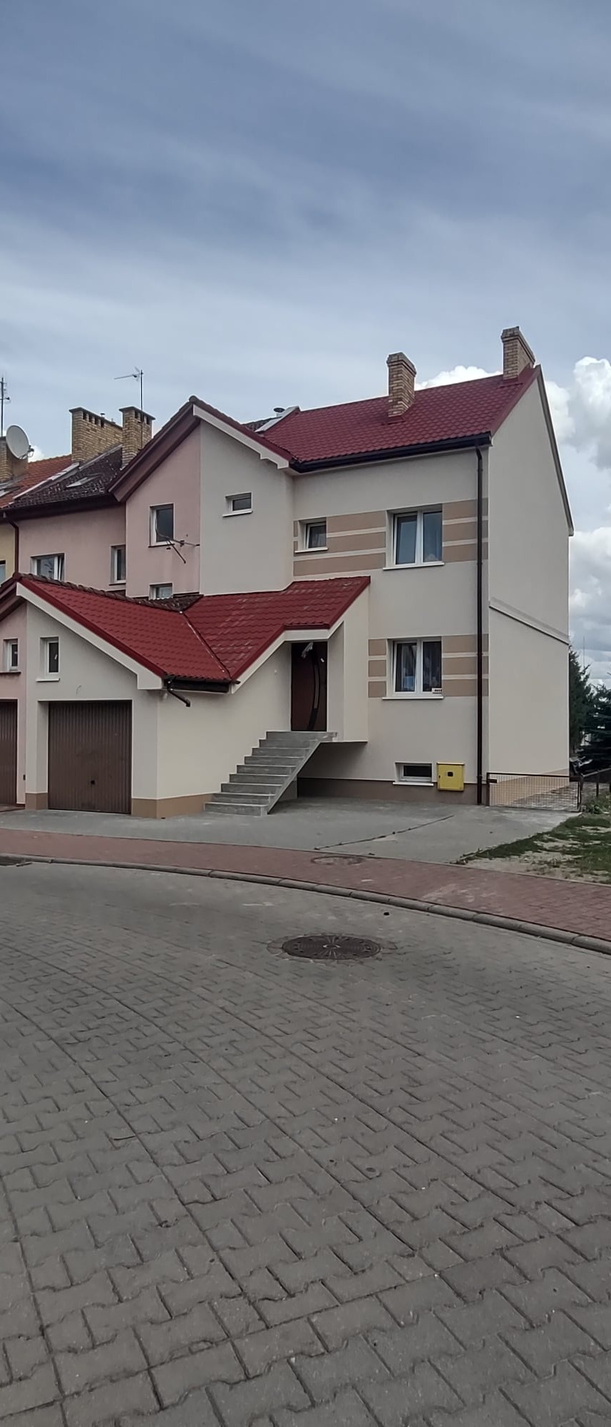 Dwupiętrowy dom szeregowy po renowacji z nową elewacją w odcieniach beżu i brązu, z czerwonym dachem i zewnętrznymi schodami prowadzącymi do wejścia.