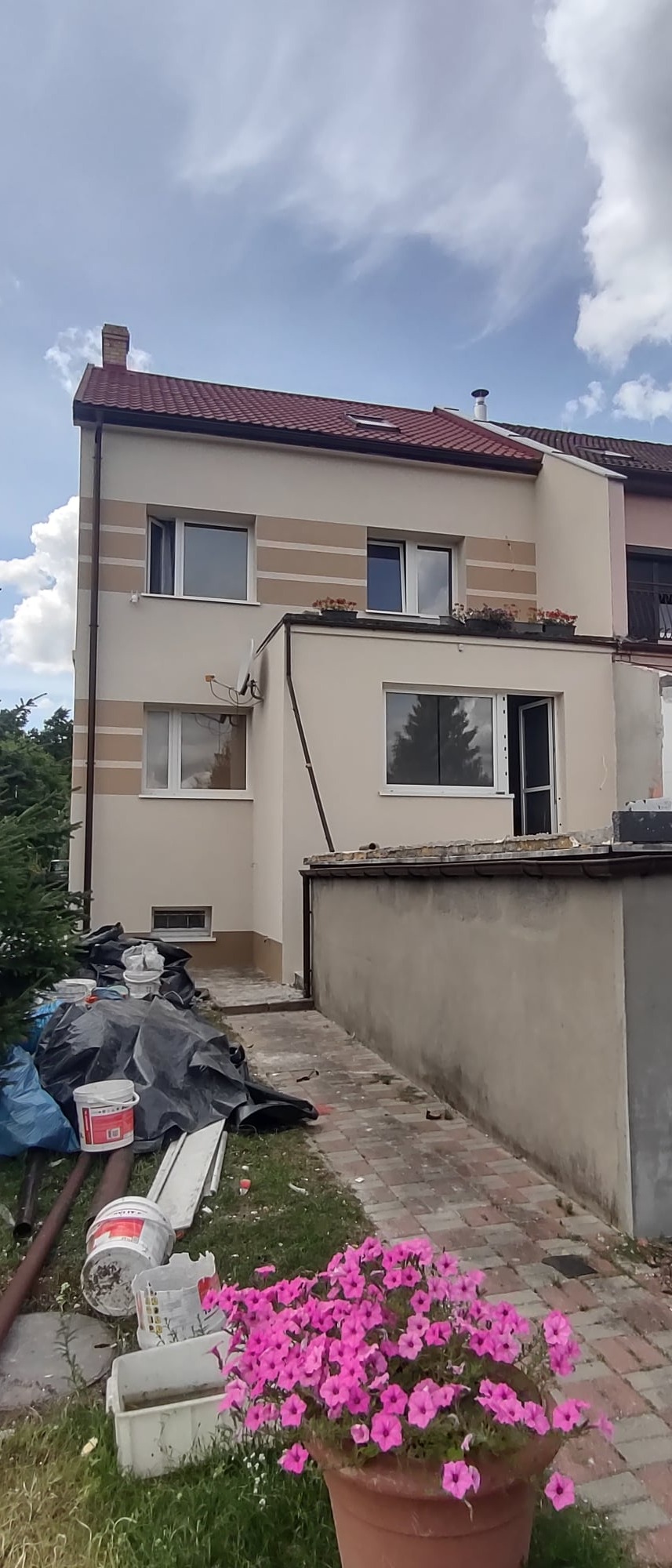 Dwupiętrowy dom z beżową elewacją i brązowymi pasami dekoracyjnymi, widoczne ślady prac malarskich, w tym puste wiadra po farbie i plandeki ochronne, donica z różowymi kwiatami na pierwszym planie.