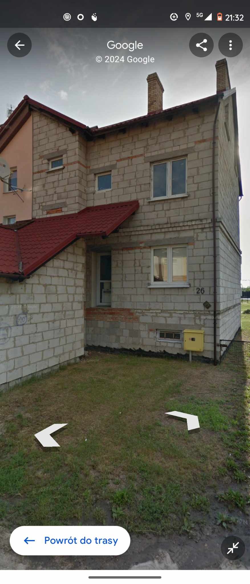 Surowy, nieotynkowany budynek z bloków, z czerwonym dachem, widoczny numer domu '26' i żółta skrzynka pocztowa, otoczony trawnikiem.