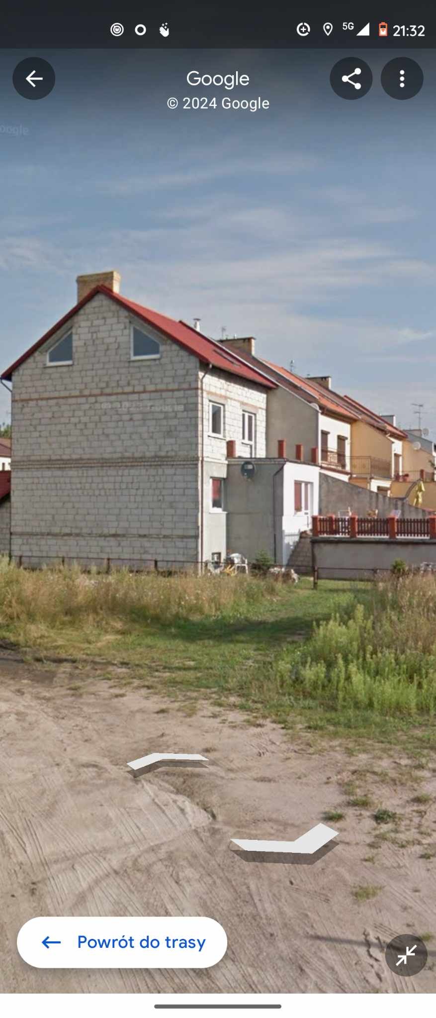 Surowy budynek w stanie deweloperskim z widoczną konstrukcją z bloczków, czerwonym dachem i małymi trójkątnymi oknami na poddaszu, otoczony niezagospodarowanym terenem z trawą.