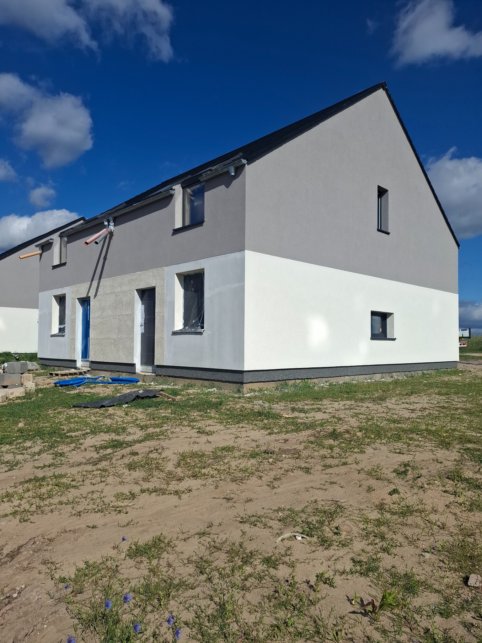 Dwukondygnacyjny budynek w trakcie prac elewacyjnych, z szarą górną częścią i białą dolną, okna zabezpieczone folią, teren wokół niezagospodarowany.