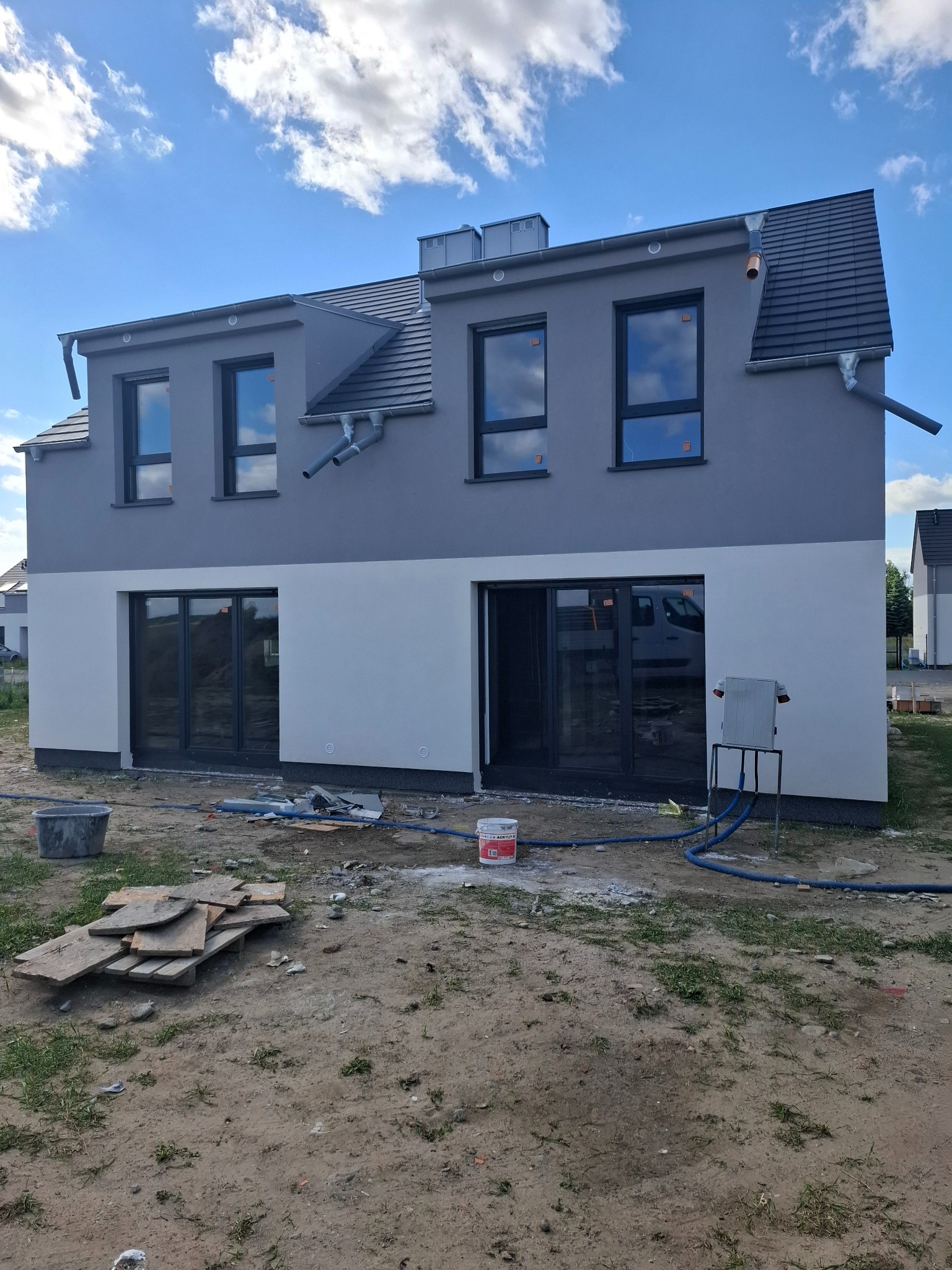 Dwukondygnacyjny dom z szarą elewacją na górze i białą na dole, z ciemnymi oknami i drzwiami balkonowymi, widoczny sprzęt do mycia elewacji i wiadro z farbą, sugerujące prace wykończeniowe.