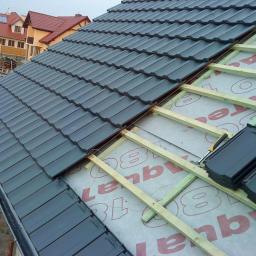 Pokryciadachowenowakowski@gmail.com - Fragment krycia dachu grafitową dachówką ceramiczną. Widoczne łaty drewniane i membrana dachowa. W tle zabudowa jednorodzinna.