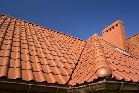 Czerwona dachówka ceramiczna na skośnym dachu domu, z widocznym kominem i systemem rynnowym w słoneczny dzień na tle błękitnego nieba.