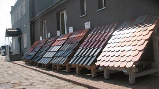 Ekspozycja różnorodnych pokryć dachowych, prezentująca szeroką gamę kolorów i wzorów dachówek, ustawiona przed budynkiem z szarą elewacją i białymi oknami.