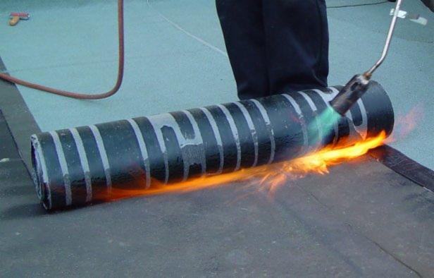 Prace dekarskie: papa termozgrzewalna zgrzewana palnikiem na dachu. Widoczny płomień, rolka papy z szarymi paskami i fragment dachu.