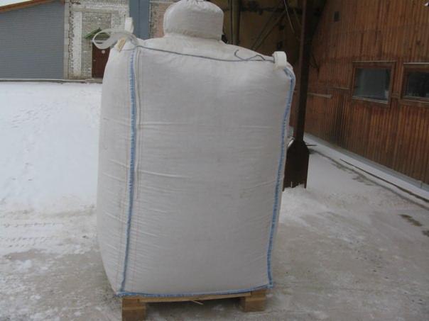 Duży, biały worek typu Big Bag na drewnianej palecie, wypełniony materiałem opałowym, na zewnątrz w zimowej scenerii. Widoczne budynki w tle.