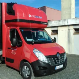 Czerwony bus Renault Master z zabudową kontenerową i logo firmy Miltrans, zaparkowany na tle budynku. Widoczna tablica rejestracyjna, antena CB i detale graficzne na plandece.
