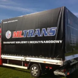 Czerwony bus dostawczy MLTRANS z czarną plandeką na tle zielonej trawy i błękitnego nieba. Widoczny napis 'Transport krajowy i międzynarodowy'.
