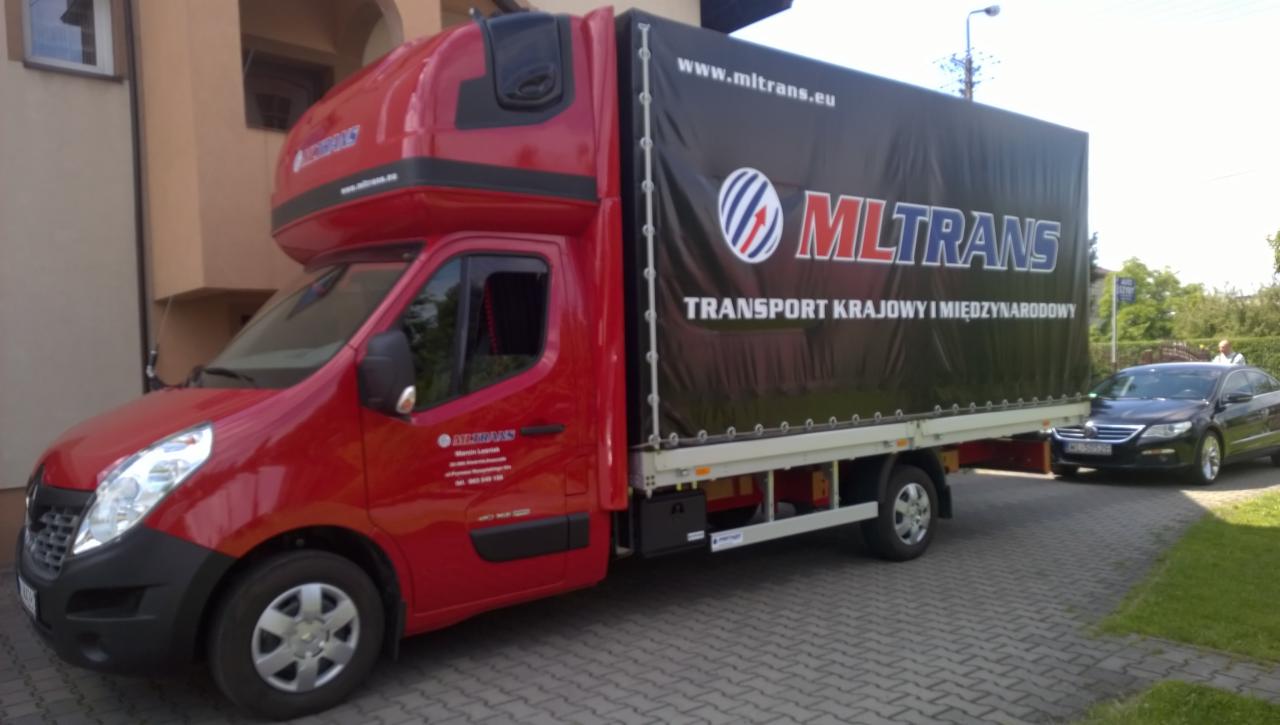 Czerwony bus transportowy Mltans z czarną plandeką, logo i adresem strony, zaparkowany na brukowanej drodze z zielenią w tle i czarnym samochodem obok.