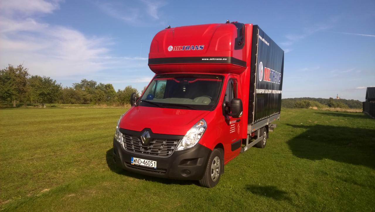 Czerwony bus Renault Master z plandeką MLTRANS na zielonym trawniku, błękitne niebo z chmurami w tle. Widoczny numer rejestracyjny WND 46051.