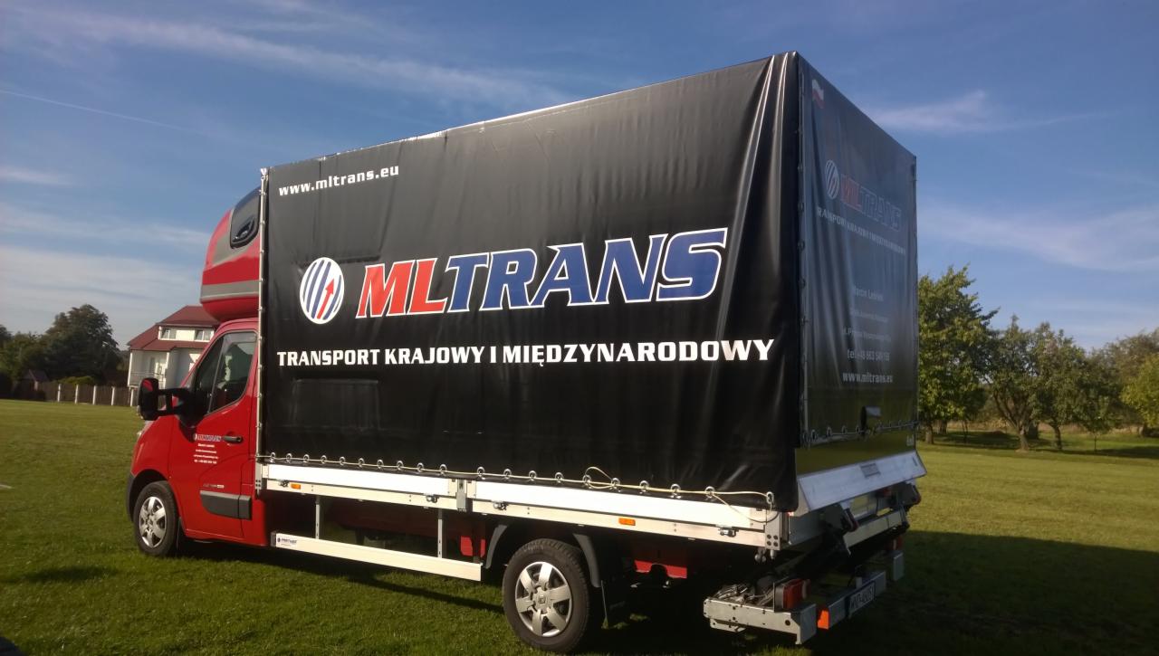Czerwony bus dostawczy MLTRANS z czarną plandeką na tle zielonej trawy i błękitnego nieba. Widoczny napis 'Transport krajowy i międzynarodowy'.