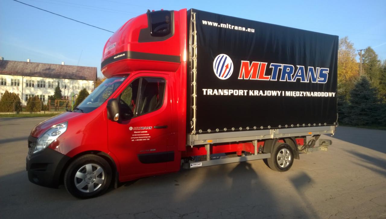 Czerwony bus dostawczy z czarną plandeką MLTRANS, transport krajowy i międzynarodowy, zaparkowany na zewnątrz w słoneczny dzień, budynek w tle.